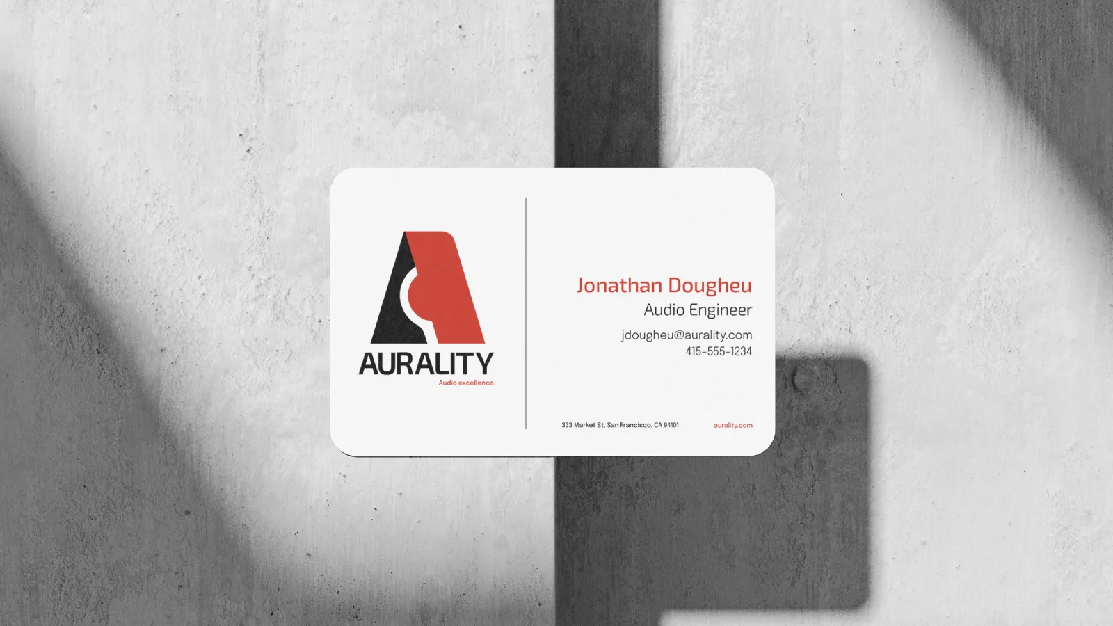 An Aurality business card on a concrete surface in diagonal light: the stacked AURALITY lockup with ‘Jonathan Dougheu, Audio Engineer’, an email and a phone number.