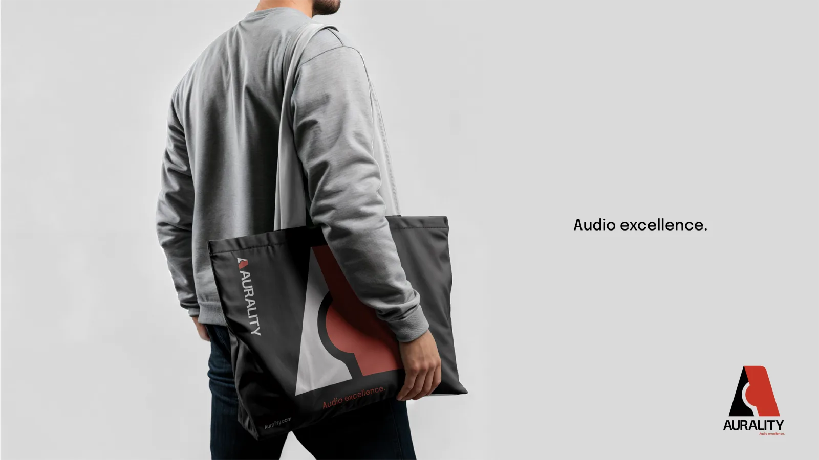 A man in a gray sweater carrying a black Aurality canvas tote printed with the red-and-black ‘A’ mark, with ‘Audio excellence.’ set to the right.