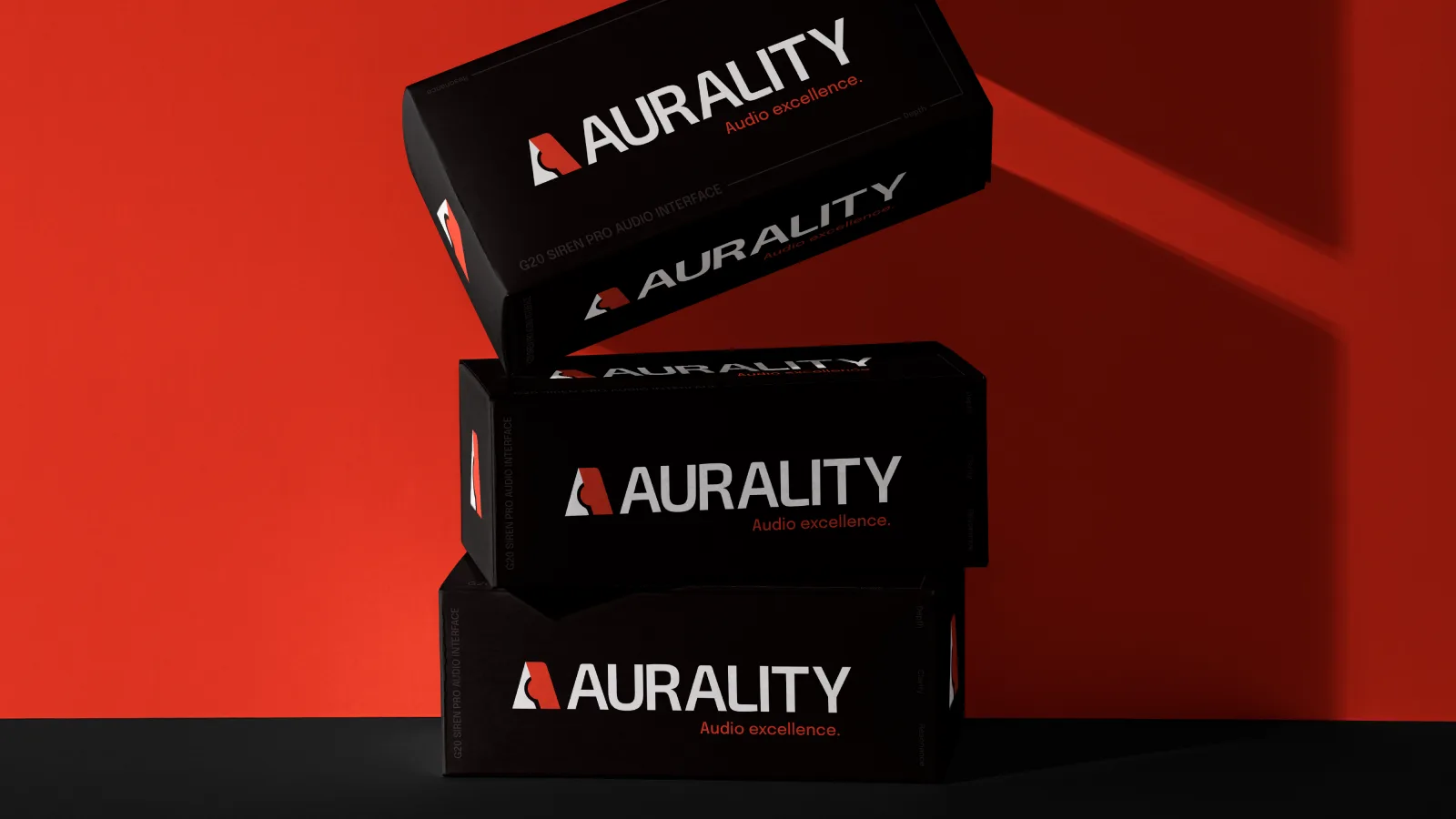 Three black Aurality product boxes stacked on a red-and-black ground with dramatic rim light, each carrying the white AURALITY lockup and ‘Audio excellence.’