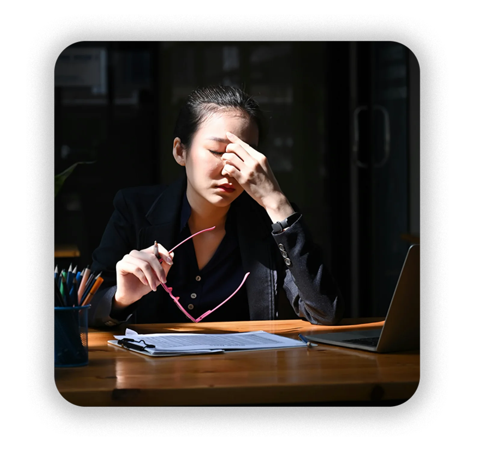 The cost, made human: a woman at a dark desk rubbing her eyes, glasses in hand, laptop and papers in front of her, struggling to hold focus through an unstructured day.