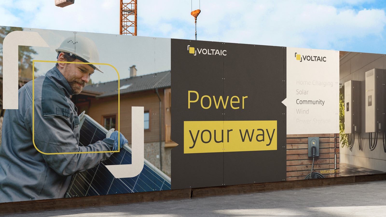 A branded Voltaic construction-site billboard under a crane: a large ‘Power your way’ board with the VOLTAIC lockup, a secondary ‘Clean Energy / Solar / Community / Wind’ panel, and a wall-charger photo at the right.